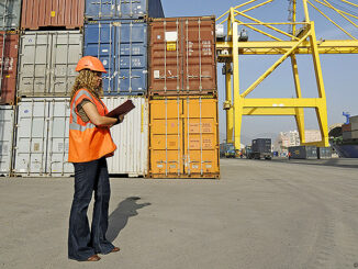 Worker with clipboard in cargo container terminal. Photo: Chris Sattlberger/Blend Images LLC
