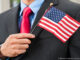 Man in a business suit holding a small American flag against his chest