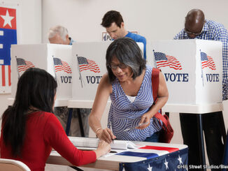 Three-quarter-length view of an Asian woman signing in to vote at a polling location, four men at voting booths behind her. MHE World.