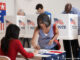 Three-quarter-length view of an Asian woman signing in to vote at a polling location, four men at voting booths behind her. MHE World.