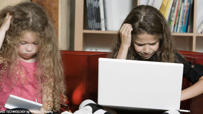 BAAYP0 Full-length view of two confused pre-school age girls sitting side by side in a living room. Each girl is looking down and stratching her head while using a wireless computer device.© PEOPLE AND TECHNOLOGY by VISION / Alamy.World.