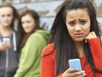 Selective focus waist-length view of a teenage girl holding her cell phone and looking at the camera with a worried or scared expression, two out-of-focus other girls looking at her in the background. © SpeedKingz/Shutterstock.com. MHE World.