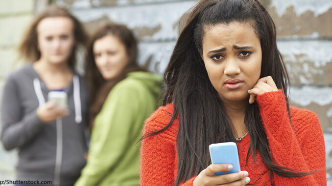 216281665 Selective focus waist-length view of a teenage girl holding her cell phone and looking at the camera with a worried or scared expression, two out-of-focus other girls looking at her in the background. © SpeedKingz/Shutterstock.com. MHE World.