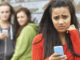 Selective focus waist-length view of a teenage girl holding her cell phone and looking at the camera with a worried or scared expression, two out-of-focus other girls looking at her in the background. © SpeedKingz/Shutterstock.com. MHE World.