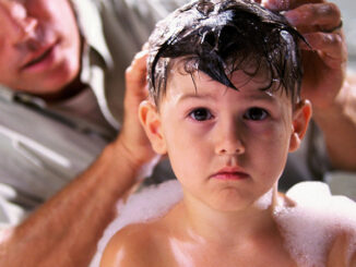 Head-and-shoulders view of a young boy in the bathtub, looking at the camera with a worried expression as his concerned father tries to wash his hair.