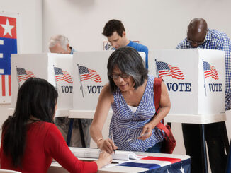 Three-quarter-length view of an Asian woman signing in to vote at a polling location, four men at voting booths behind her.