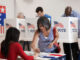 Three-quarter-length view of an Asian woman signing in to vote at a polling location, four men at voting booths behind her.