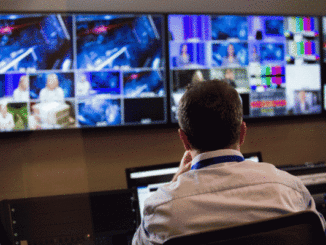Head and shoulders, selective focus back view of a male TV director sitting at a vision mixing panel in a television studio with flat panel monitors displaying various filmed scenes in the background