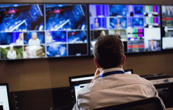062817_02_IS Head and shoulders, selective focus back view of a male TV director sitting at a vision mixing panel in a television studio with flat panel monitors displaying various filmed scenes in the background