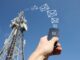 Man's hand holding a smart phone in front of telecommunication mast under a blue sky