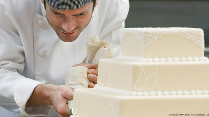 061318_01P_FS A man carefully decorating a wedding cake with lace icing details.
