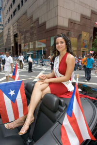Miss Teen Princess, an hispanic female wearing a formal dress, waves a Puerto Rican flag while riding in a convertible car in a Puerto Rico Parade in New York City