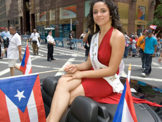 Miss Teen Princess, an hispanic female wearing a formal dress, waves a Puerto Rican flag while riding in a convertible car in a Puerto Rico Parade in New York City