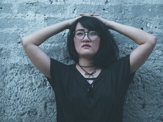 Waist-up view of an Asian girl with her hands on top of her head leaning against a cement wall