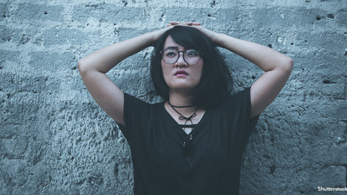Fear, loneliness, depression, abuse, addiction Waist-up view of an Asian girl with her hands on top of her head leaning against a cement wall