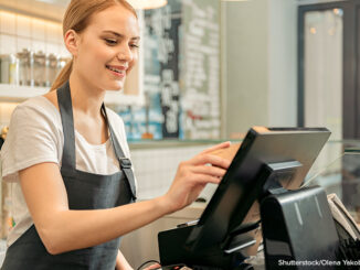 Waist-up, cropped selective focus view of a smiling young woman standing behind a counter and using a touchscreen to process payment in a coffee shop or cafe