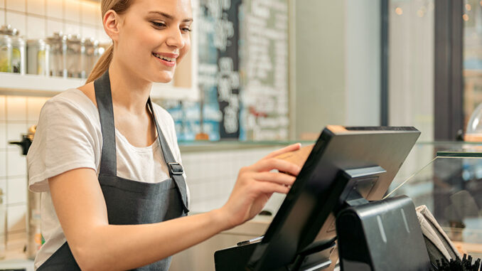 Cheerful shop assistant using digital device for payment Waist-up, cropped selective focus view of a smiling young woman standing behind a counter and using a touchscreen to process payment in a coffee shop or cafe