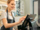 Waist-up, cropped selective focus view of a smiling young woman standing behind a counter and using a touchscreen to process payment in a coffee shop or cafe
