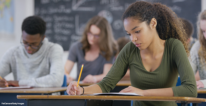 A mixed race teenage girl is taking a high school standardized test in class. She looks down and works on the test.