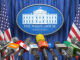 Media microphones set up at podium with White House Washington sign on curtained background, flanked by American flagss