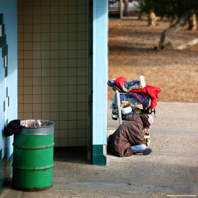 A homeless person sitting on a sidewalk near their possessions in a shopping cart.