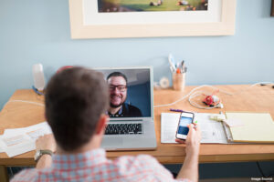 man sitting at a desk, holding a cell phone and videoconferencing with another man on his laptop