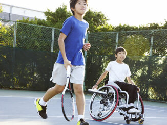 A teenage boy running with another teenage boy in a wheelchair on a tennis court