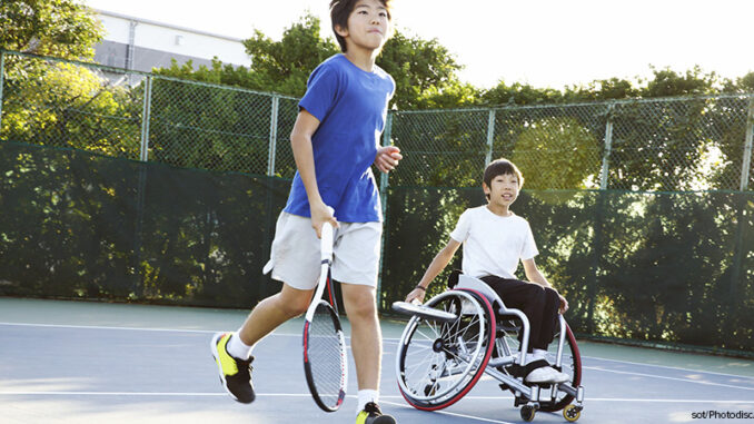 A teenage boy running with another teenage boy in a wheelchair on a tennis court A teenage boy running with another teenage boy in a wheelchair on a tennis court