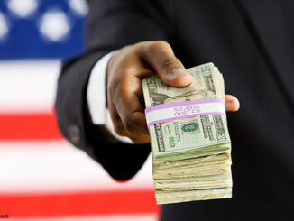 Suited political figure standing in front of American flag background, handing a bundled stack of dollar bills to the viewer