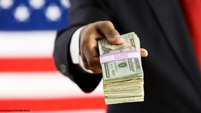 101220_01P_FS Suited political figure standing in front of American flag background, handing a bundled stack of dollar bills to the viewer