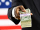 Suited political figure standing in front of American flag background, handing a bundled stack of dollar bills to the viewer