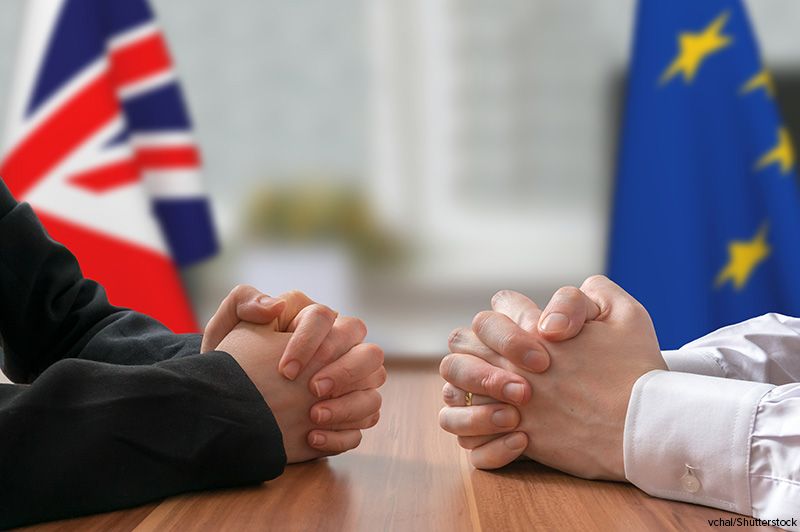 Image of people's hands clasped at a table with the flags of Great Britain and the European Union in the background