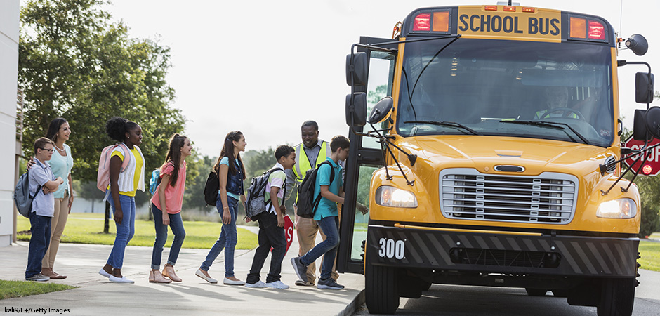 A group of six middle school students, with their teacher and a school crossing guard, boarding a yellow school bus.