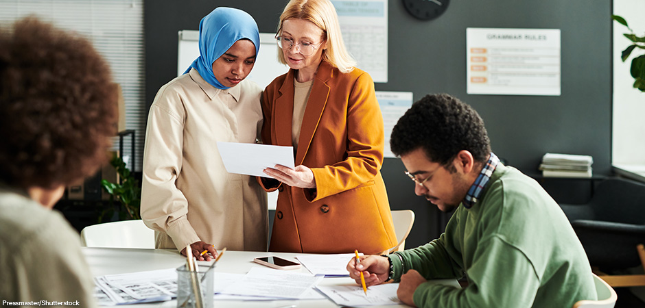 Blonde woman showing document to Muslim female student in hijab while both standing by workplace
