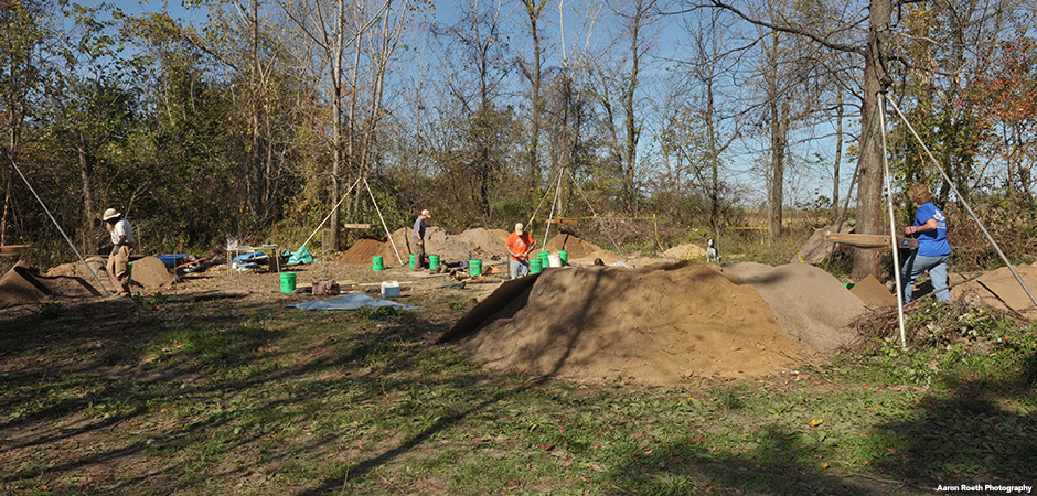 Wide view of working archaeologists digging, walking and using a tripod sifting box at the site of an archaeological dig