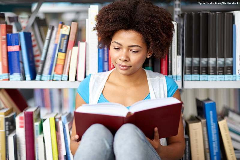 African American female student reading book sitting on floor at library