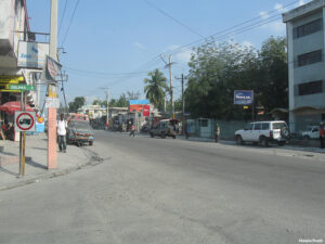Wide view of a city intersection showing run-down buildings, old cars and people walking on the sidewalks and street in Haiti.