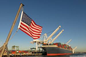 An American flag waves in the foreground at this US port, where a cargo ship loaded with containers is berthed beneath giant cranes on a clear blue sky day. 