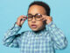 Portrait of little black boy wearing eyeglasses looking at camera, standing on blue studio background.